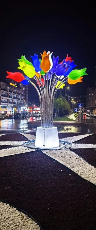 Gece, çakıllı peyzaj adasında aydınlatmalı beyaz kaideden yükselen renkli lale buketi biçimli ışıklı heykel