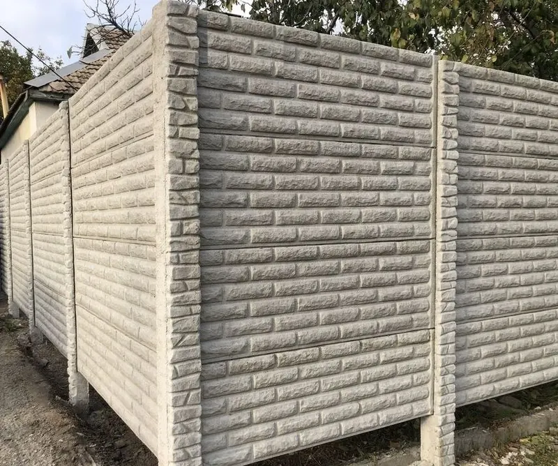 Sokak kenarında köşe dönen, kabartmalı tuğla desenli açık gri beton çit panelleri ve aynı dokulu sütunlar